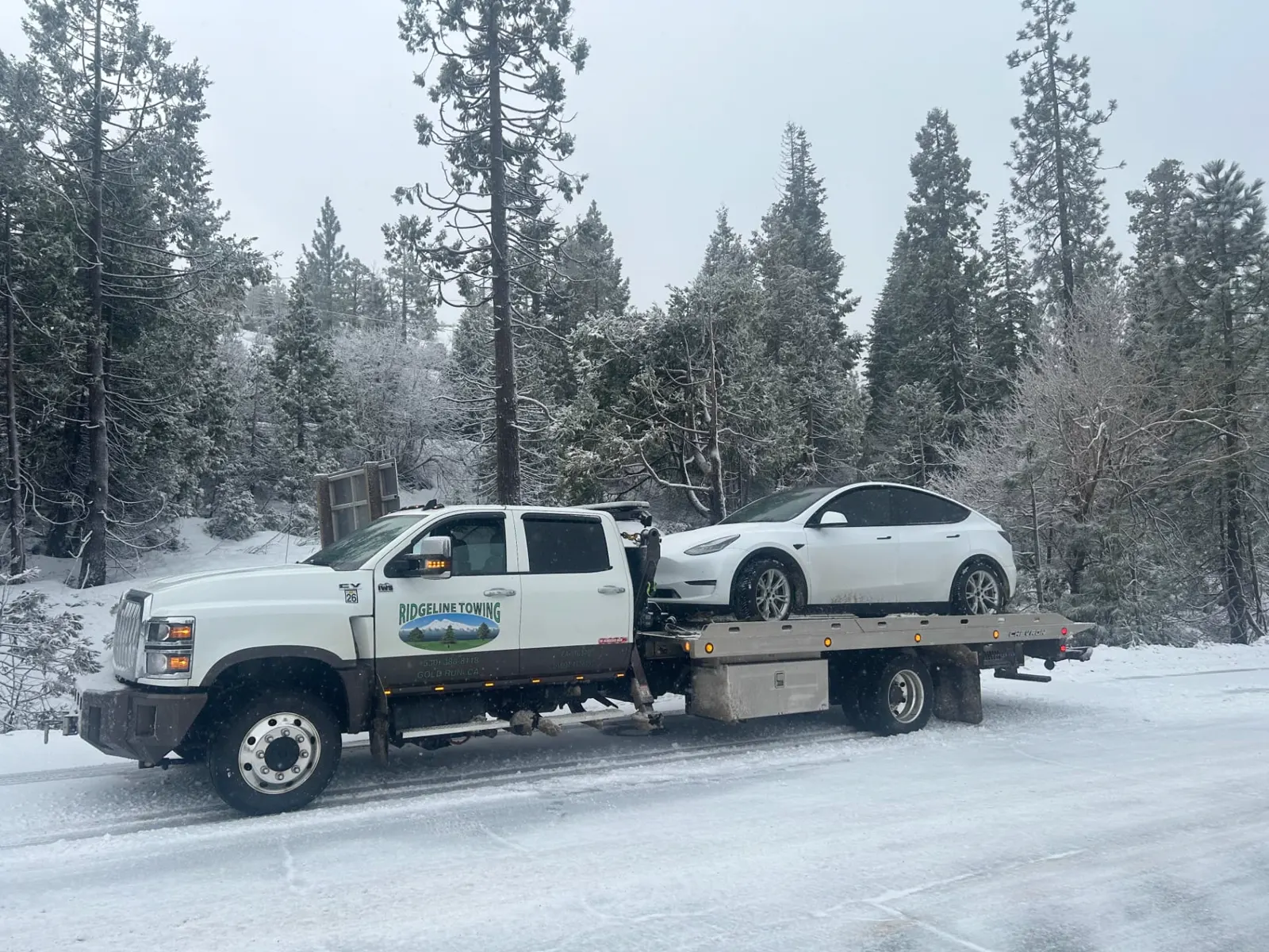 Ridgeline Towing flatbed hauling a white Tesla through falling snow in the Sierra Nevada