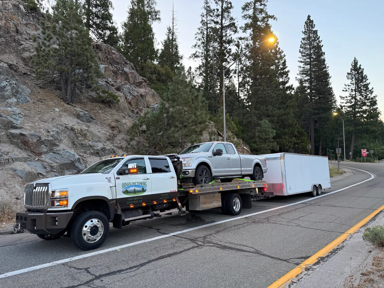 Ridgeline Towing flatbed hauling a pickup and enclosed trailer on a Sierra mountain road